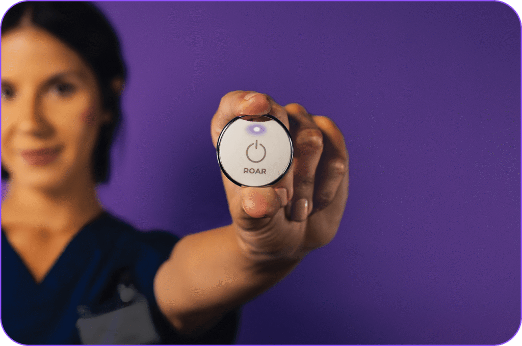 Hand holding up a ROAR wearable panic button with its light glowing purple
