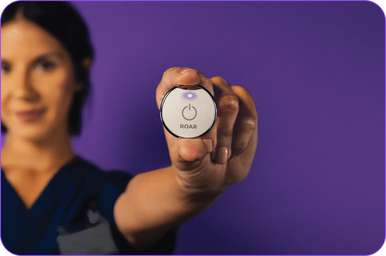 Hand holding up a ROAR wearable panic button with its light glowing purple