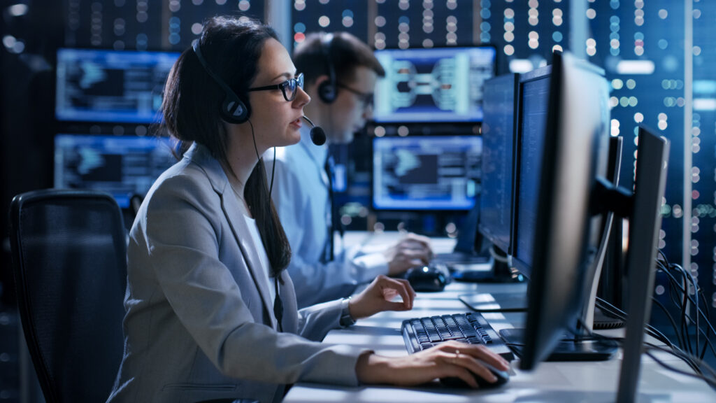 Emergency dispatchers wearing headsets working at computer monitors in a dimly lit monitoring center.