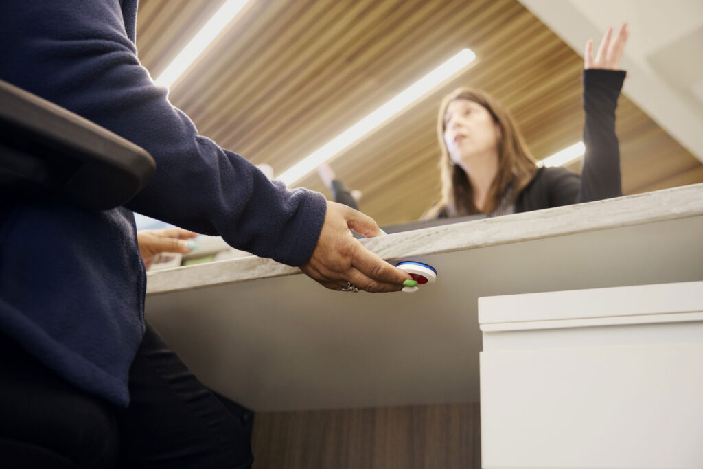 Person reaching up to press a ROAR panic button mounted discreetly underneath a counter or desk ledge.