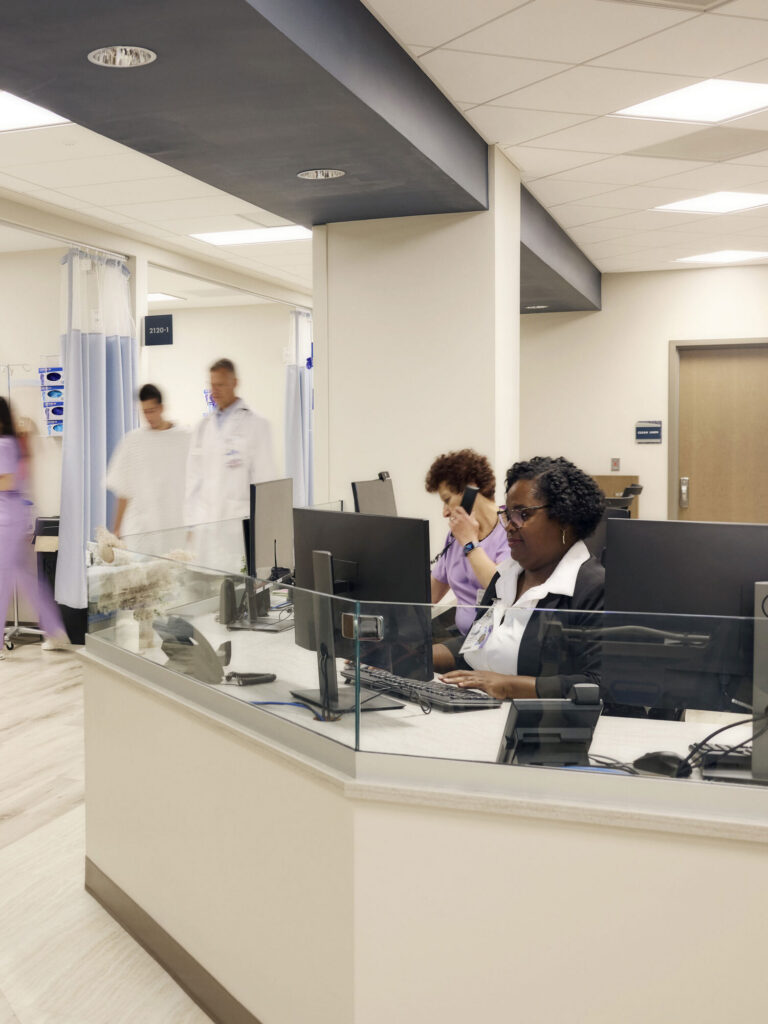 Hospital staff working at a nurses' station behind a glass partition while a patient and doctor walk past privacy curtains in the background.