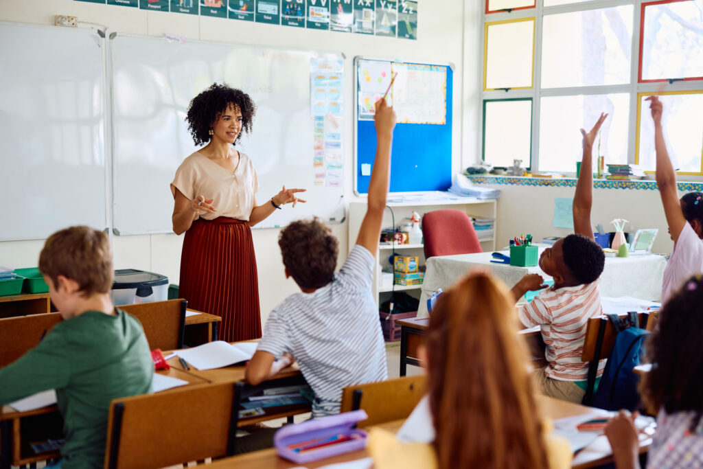 Teacher leading a lesson as a student raises their hand in a classroom.