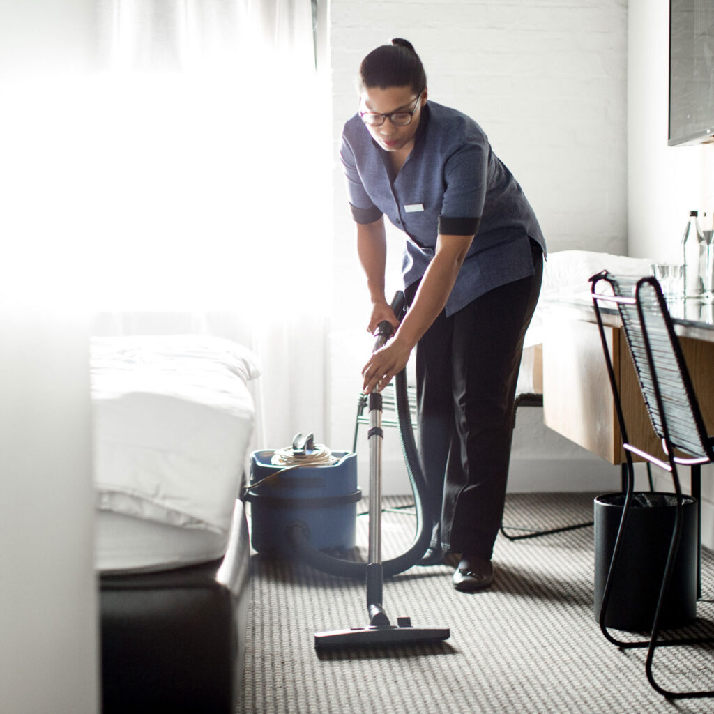 Housekeeper vacuuming a brightly lit hotel guest room.