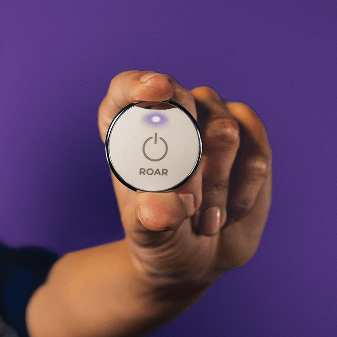 Hand holding up a ROAR wearable panic button with its light glowing purple