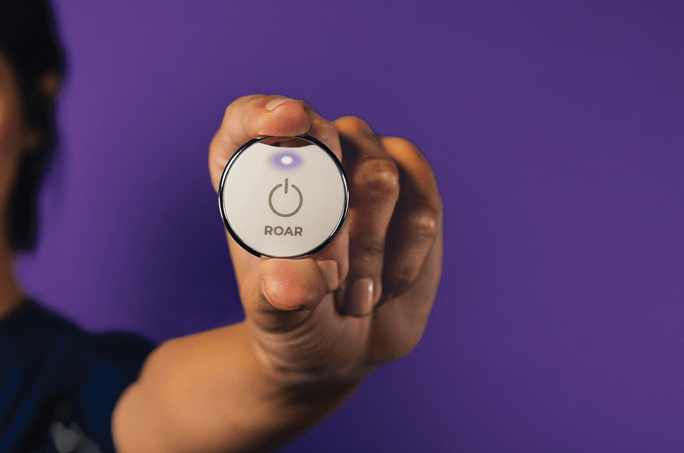 Hand holding up a ROAR wearable panic button with its light glowing purple
