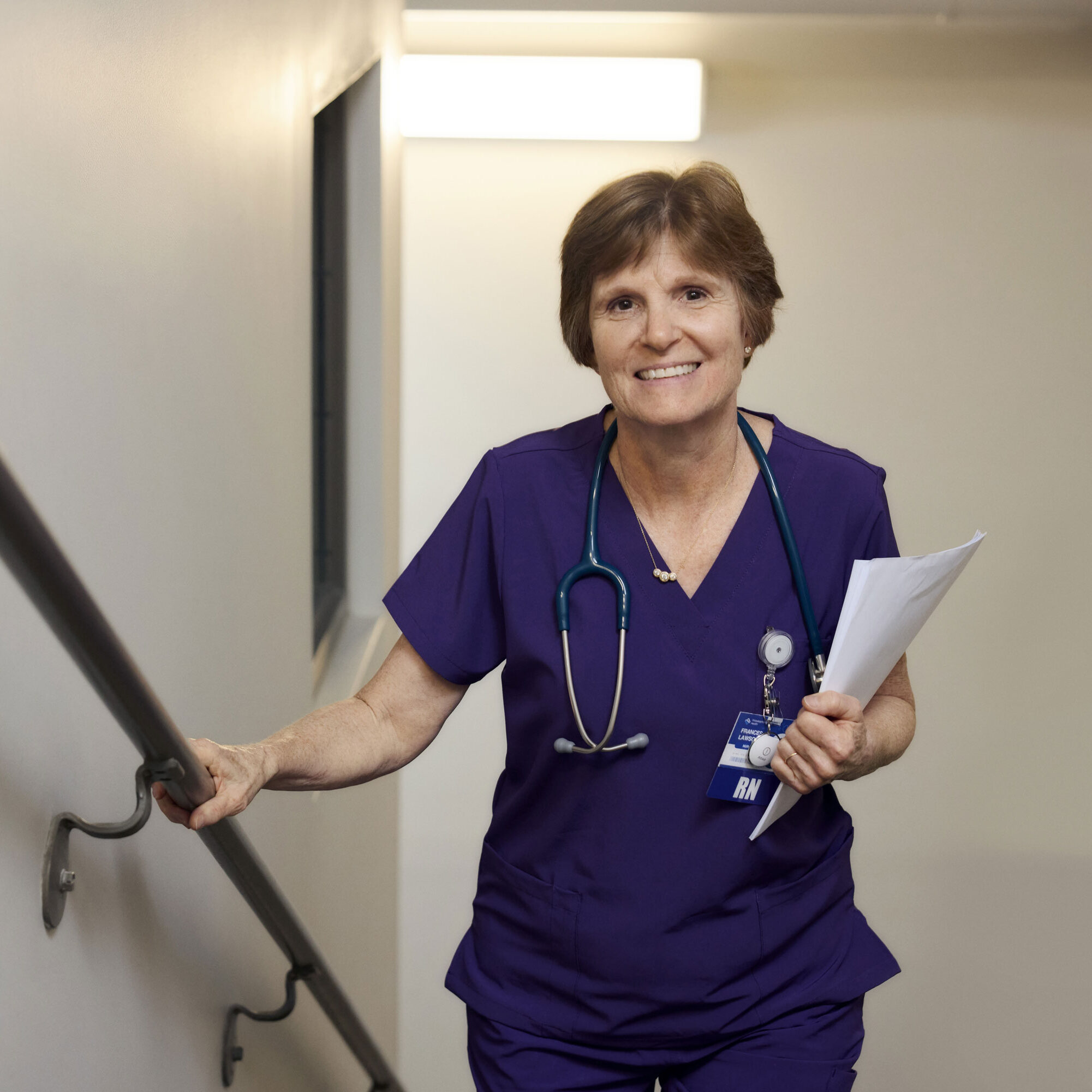 Nurse walking up a staircase holding papers and smiling, wearing a ROAR Alert Device on dark purple scrubs