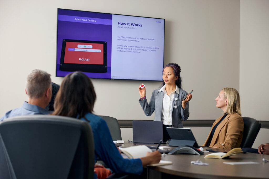 asian woman holding a ROAR panic button with a red center and a mobile phone to several people seated at a conference room table with a presentation screen in the background showing the a ROAR End User training slide